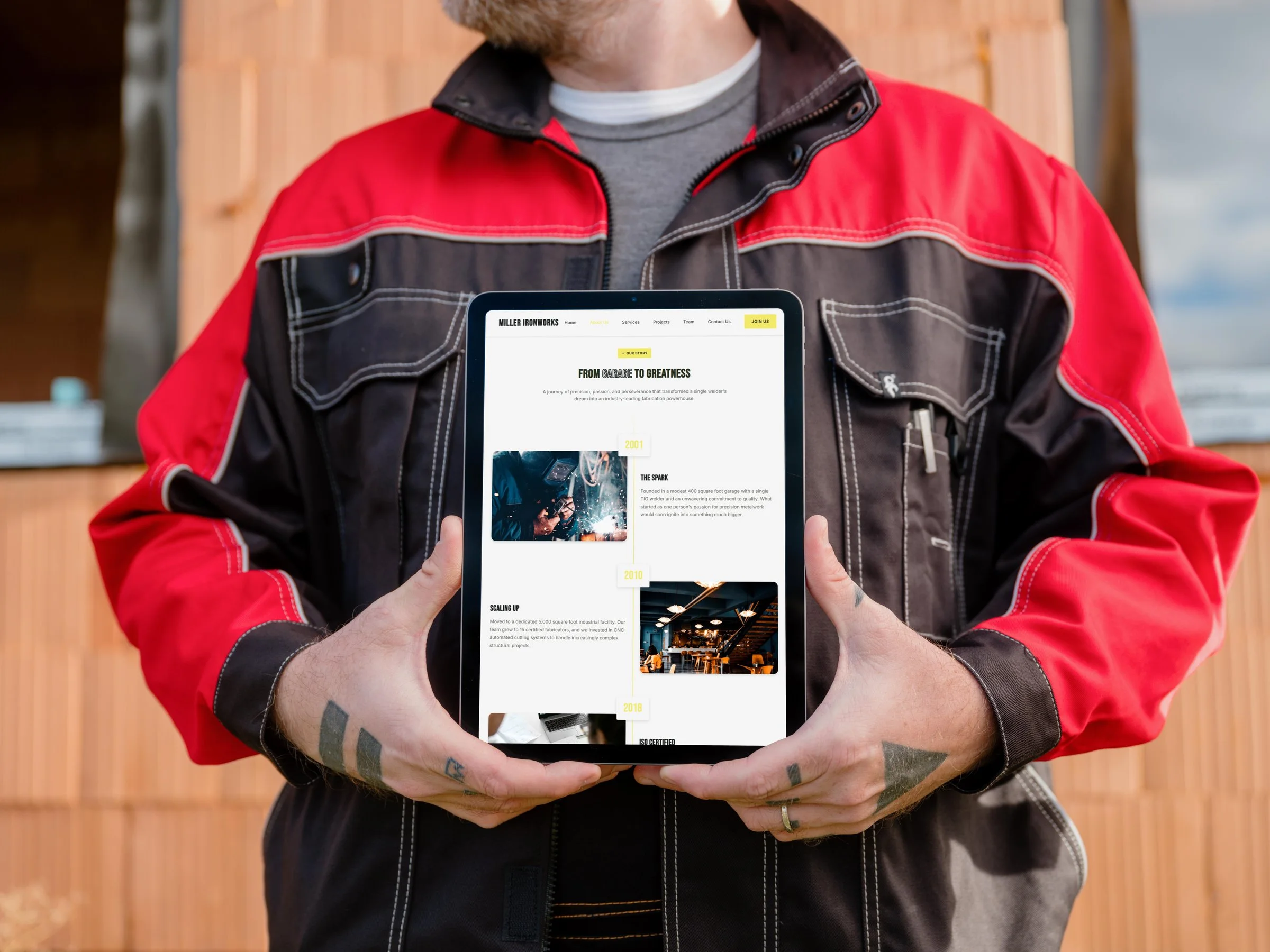 A person in a work jacket holding a tablet showing the Miller Ironworks "From Garage to Greatness" page.